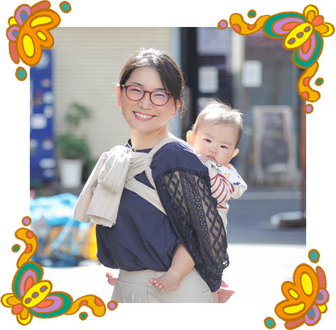 大久保久美子さんの写真。明るい昼間、ベビースリングで赤ちゃんを背負っている。